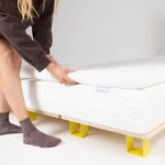 Eine Person, die graue Socken und einen dunklen Pullover trägt, verstellt eine weiße Matratzenauflage auf einer Matratze mit einem Holzrahmen und gelben Latten.