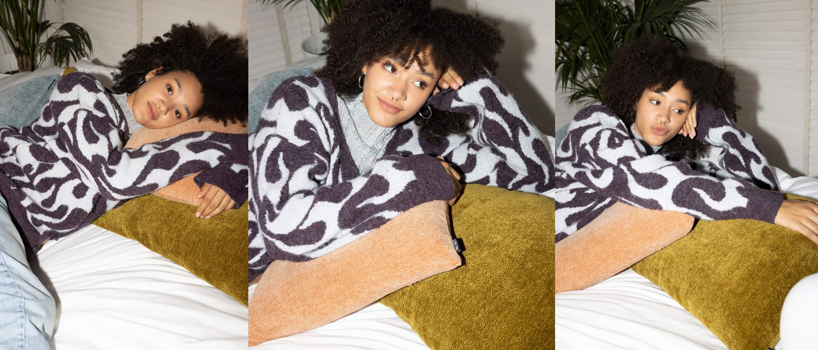 Eine junge Frau mit lockigem Haar liegt auf einem Bett und trägt einen schwarz-weiß gemusterten Pullover. Sie stützt sich auf senf- und hellbraune Kissen und blickt nachdenklich zur Seite in einem gut beleuchteten Raum mit weißen Wänden und grünen Pflanzen.