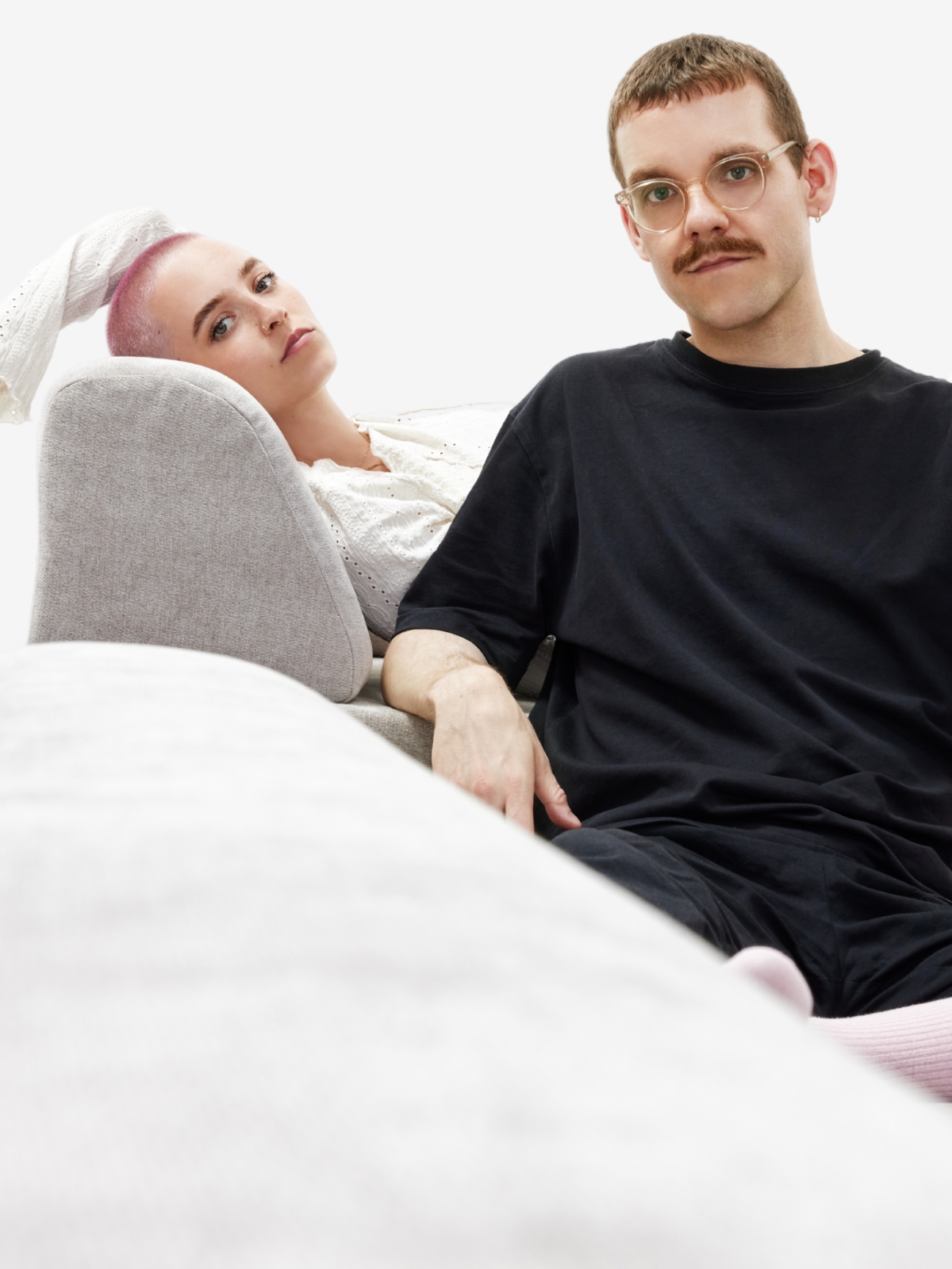 Eine Frau mit hellrosa Haaren lehnt sich auf einer hellgrauen Couch zurück und blickt in die Kamera, während ein Mann mit kurzen Haaren, Brille und Schnurrbart neben ihr sitzt, bekleidet mit einem schwarzen Hemd vor weißem Hintergrund.