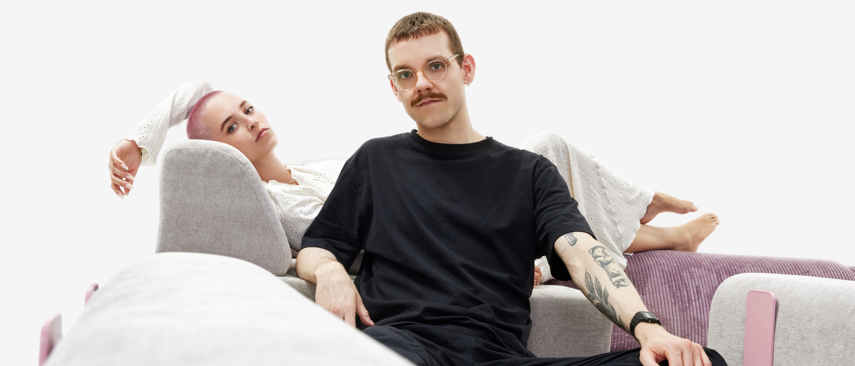 Eine Frau mit kurzen hellen Haaren lehnt sich auf einem hellen Sofa zurück, während ein Mann mit Brille, Schnurrbart und Tattoos in einem schwarzen T-Shirt und einer Hose vor ihr sitzt. Beide wirken entspannt vor einem weißen Hintergrund.