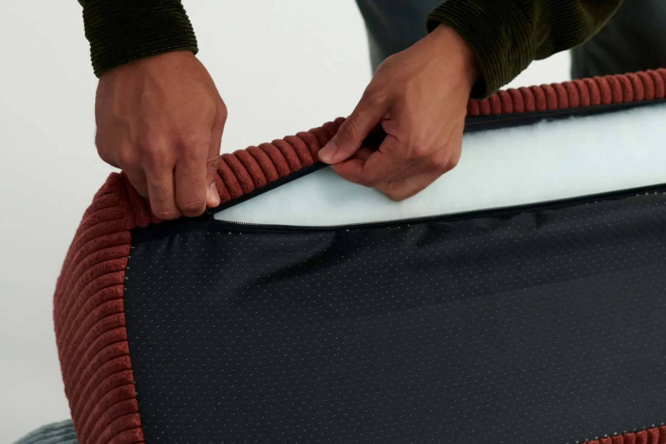 Die Hände einer Person öffnen den Reißverschluss eines dunklen, strukturierten Stoffbezugs mit rötlich-braunem Rand und geben eine weiße Innenschicht frei, die möglicherweise von einem Kissen oder einer Matratze stammt.