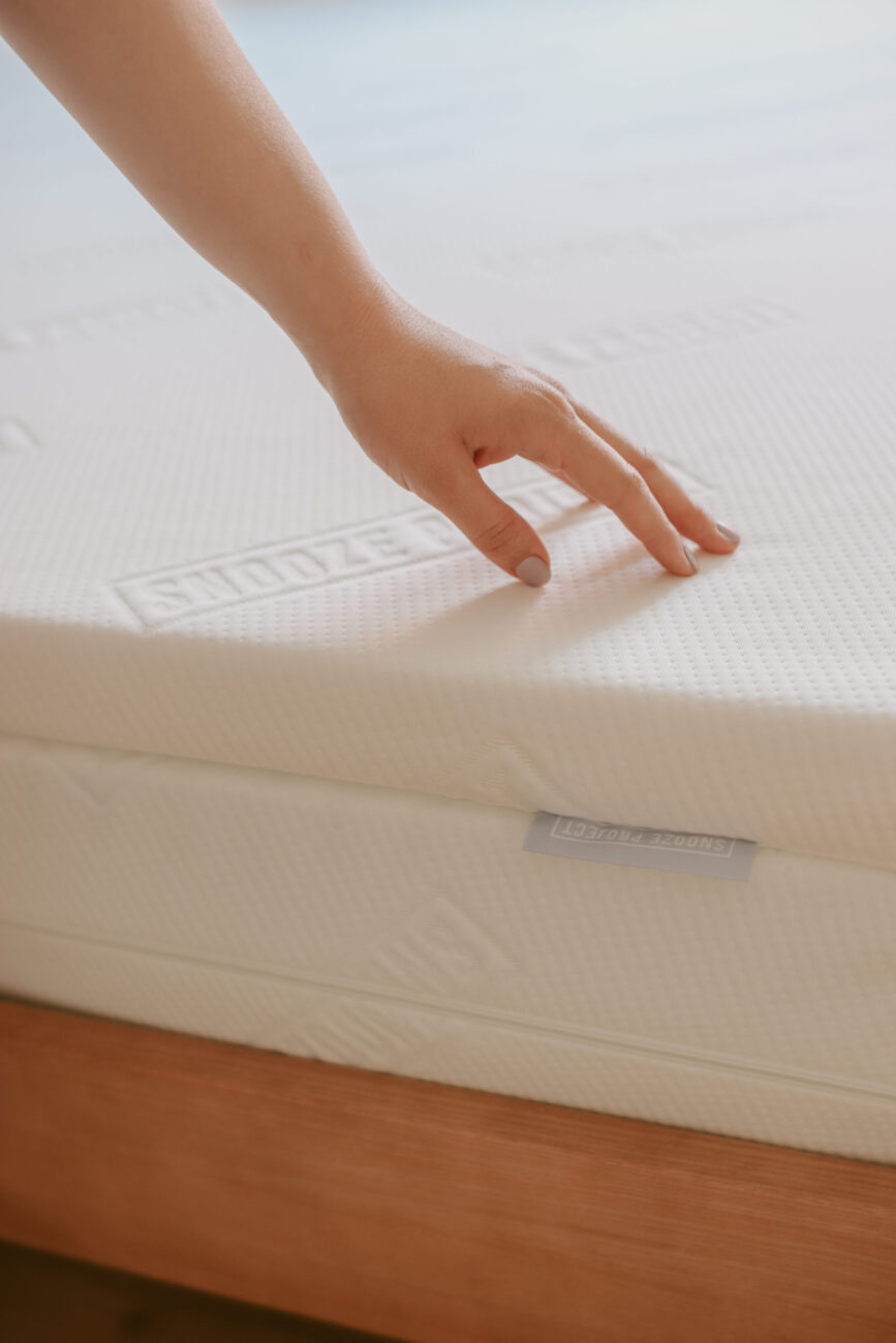 Eine Hand drückt sanft auf eine weiße, strukturierte Matratze, um ihre Weichheit zu testen. Das Etikett Topper Mads ist sichtbar, während die Matratze auf einer hölzernen Oberfläche ruht und Sie in eine Erlebniswelt des Komforts einlädt.