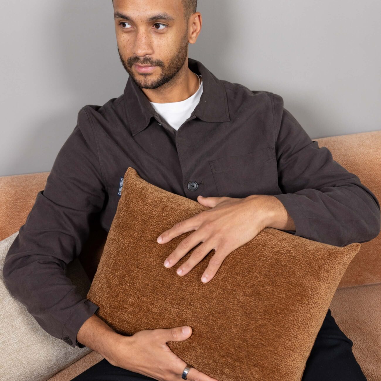 SPKDEKO45x30-Mood_img001 Ein Mann mit gestutztem Bart sitzt auf einer beigen Couch und trägt eine dunkelbraune Jacke über einem weißen Hemd. Er hält mit beiden Händen ein strukturiertes, hellbraunes Kissen über seine Brust und schaut zur Seite.