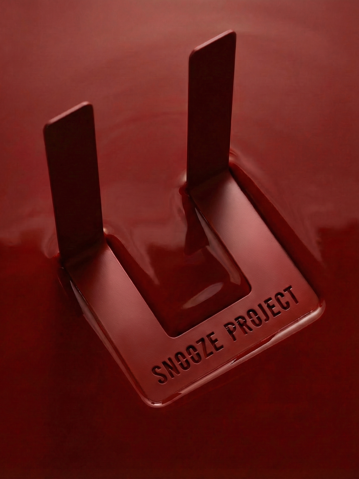 Ein metallisches, U-förmiges Objekt, auf dessen Oberfläche der Schriftzug SNOOZE PROJECT eingraviert ist, vor einem glänzenden roten Hintergrund.