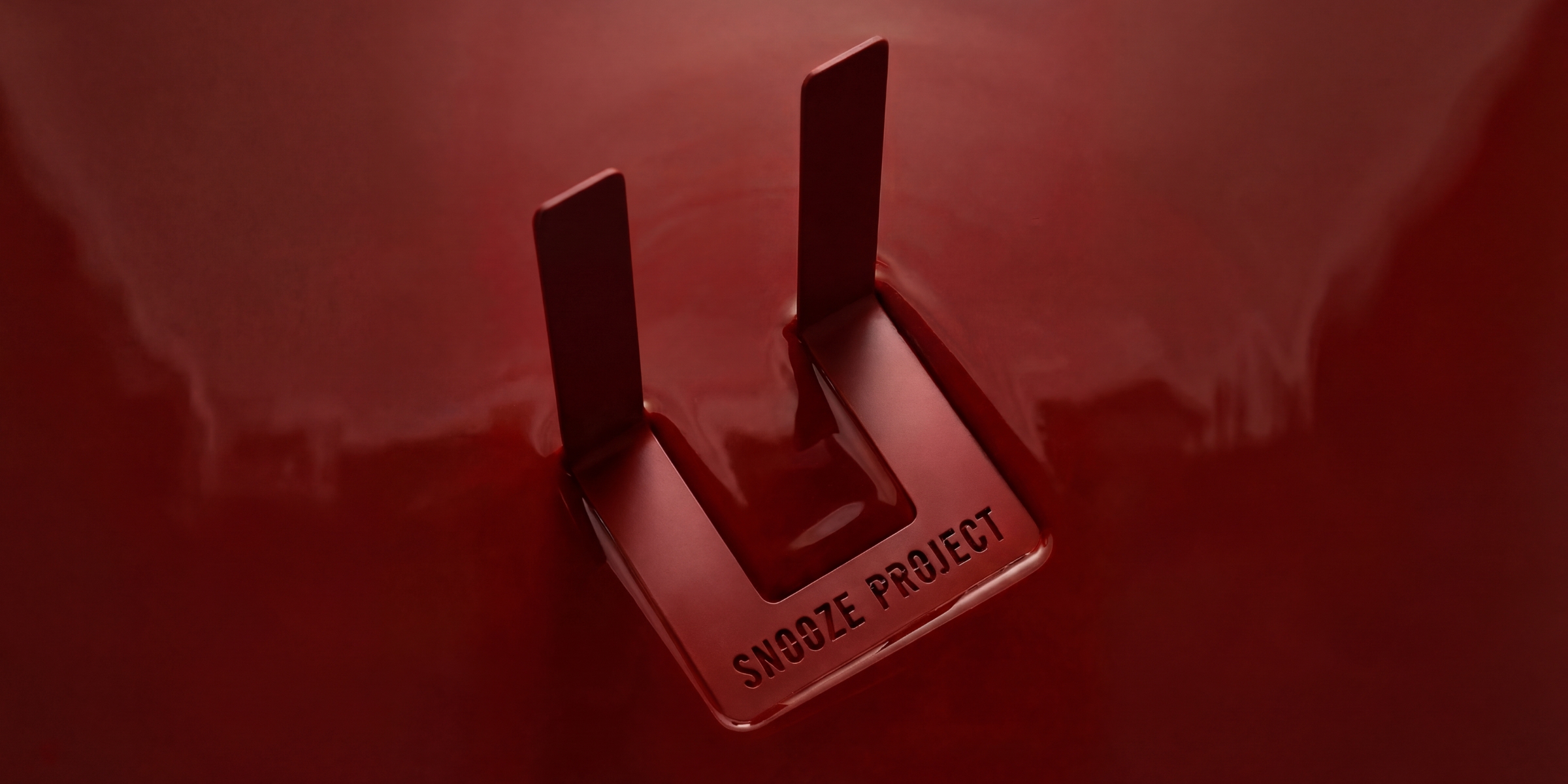 Nahaufnahme einer glänzenden roten Oberfläche mit zwei aufrecht stehenden Metallzacken, in deren Nähe die Worte SNOOZE PROJECT eingraviert sind.