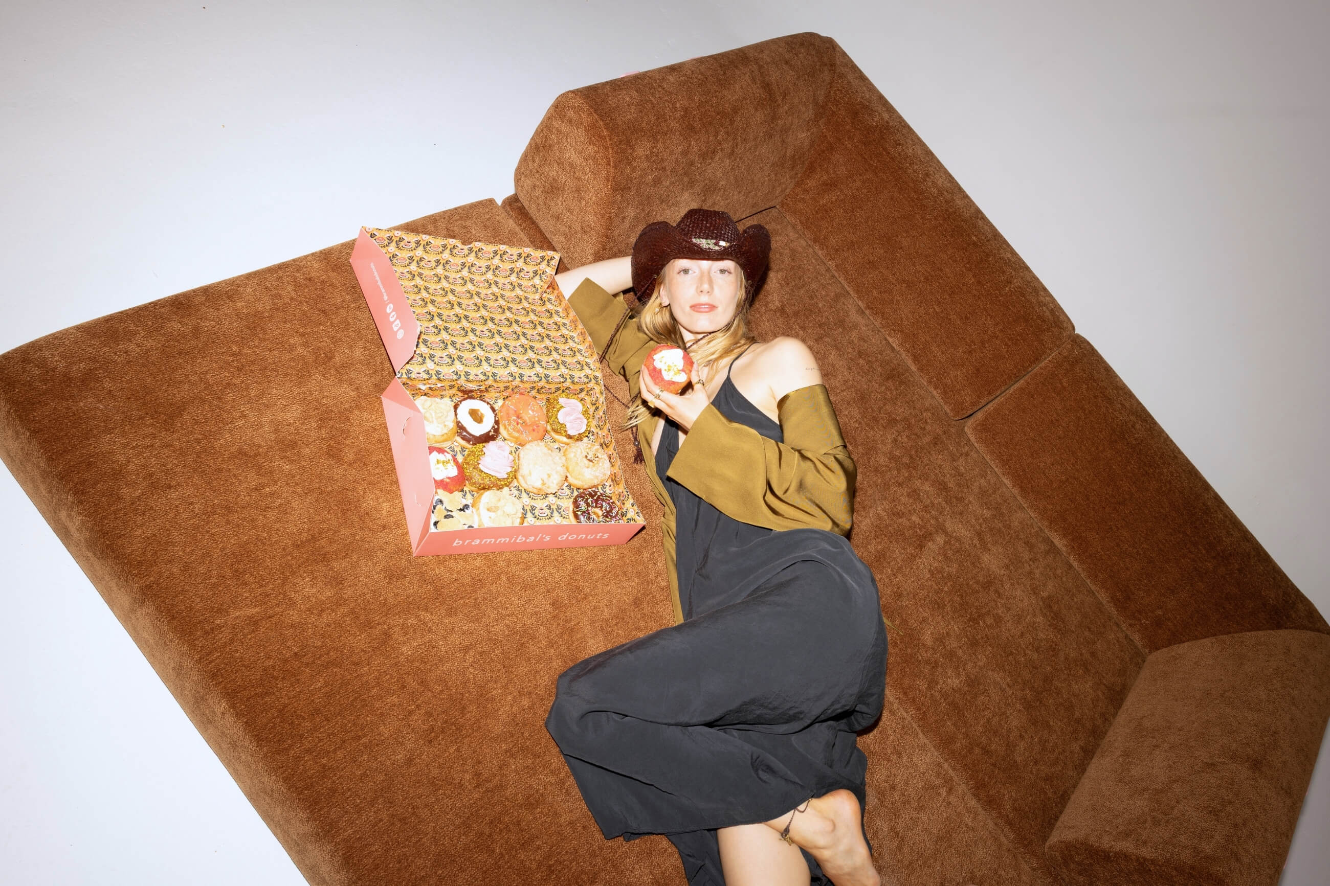 Eine Frau in einem dunklen Kleid und mit Cowboyhut liegt auf einem braunen Sofa und hält einen Donut in der Hand. Neben ihr steht eine geöffnete rosafarbene Schachtel, die mit verschiedenen Donuts gefüllt ist.