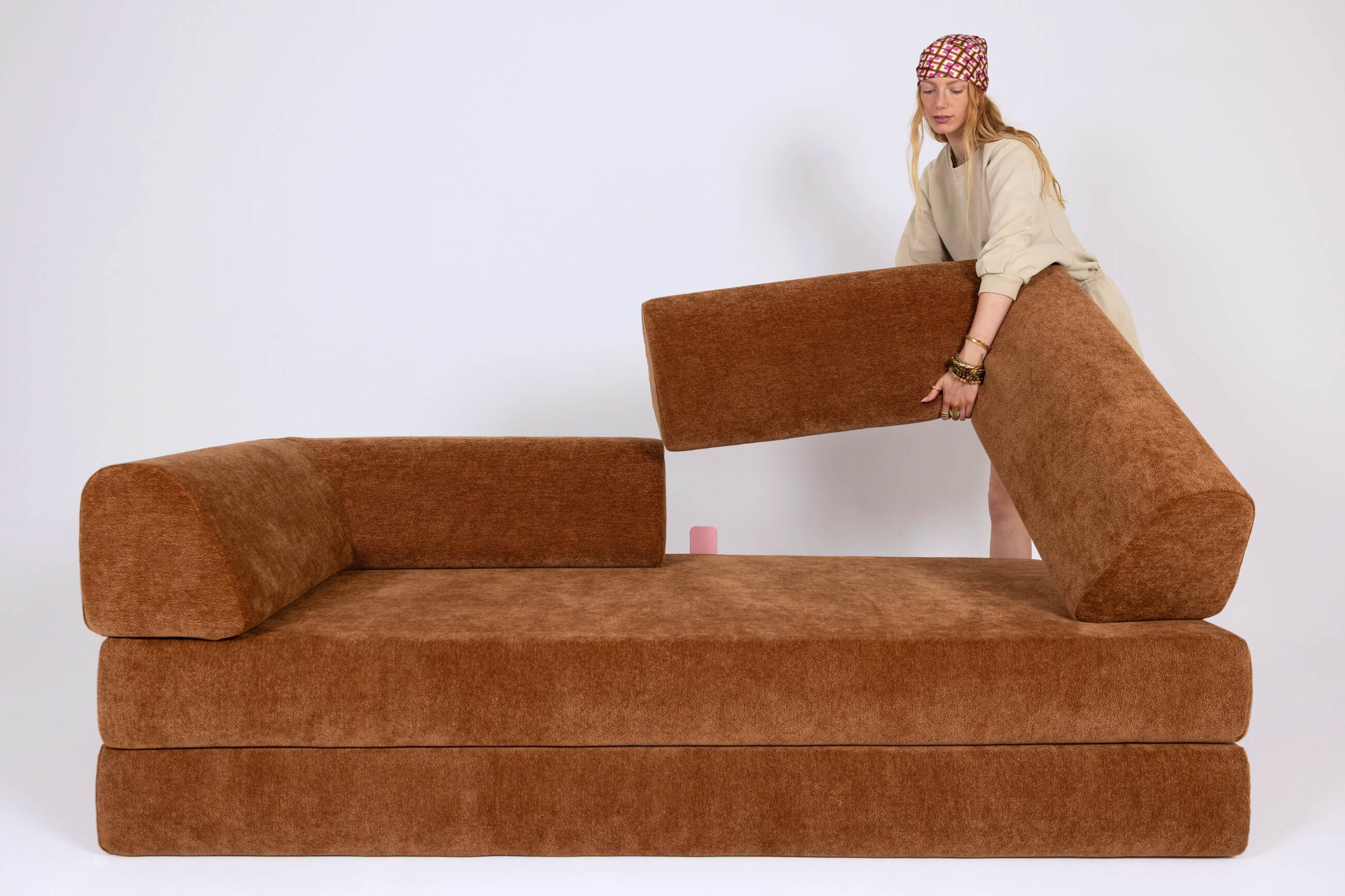 Eine Person verstellt ein großes, braunes, modulares Sofa mit abnehmbaren Kissen in einem minimalistischen, weißen Raum.