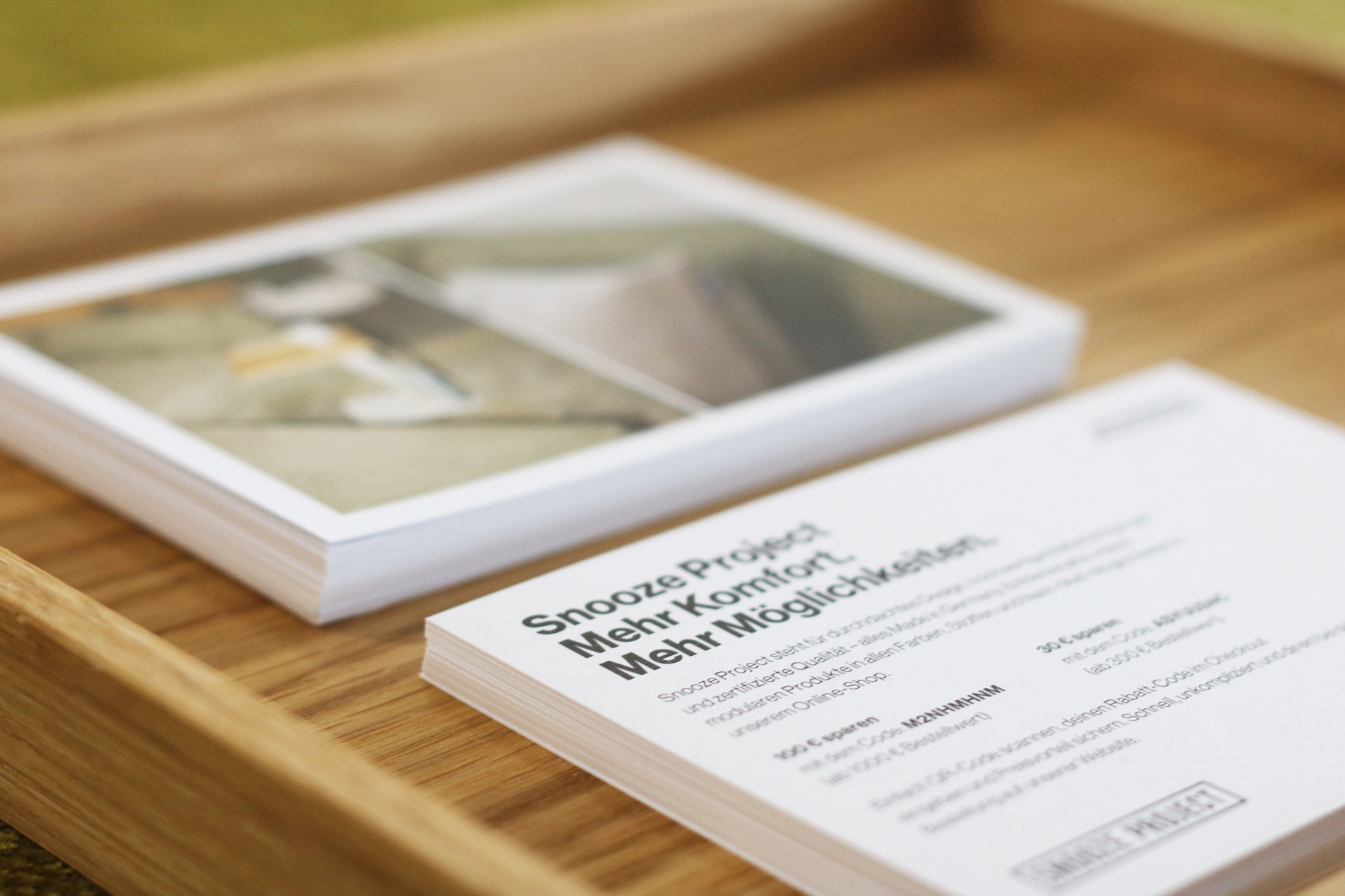 Ein Stapel Postkarten befindet sich in einem Holztablett. Die oberste Postkarte mit der Aufschrift "Snooze Project" deutet auf Verbindungen zu Friends of Snooze Project hin, während im Hintergrund ein unscharfes Bild auf einer anderen Postkarte zu sehen ist.