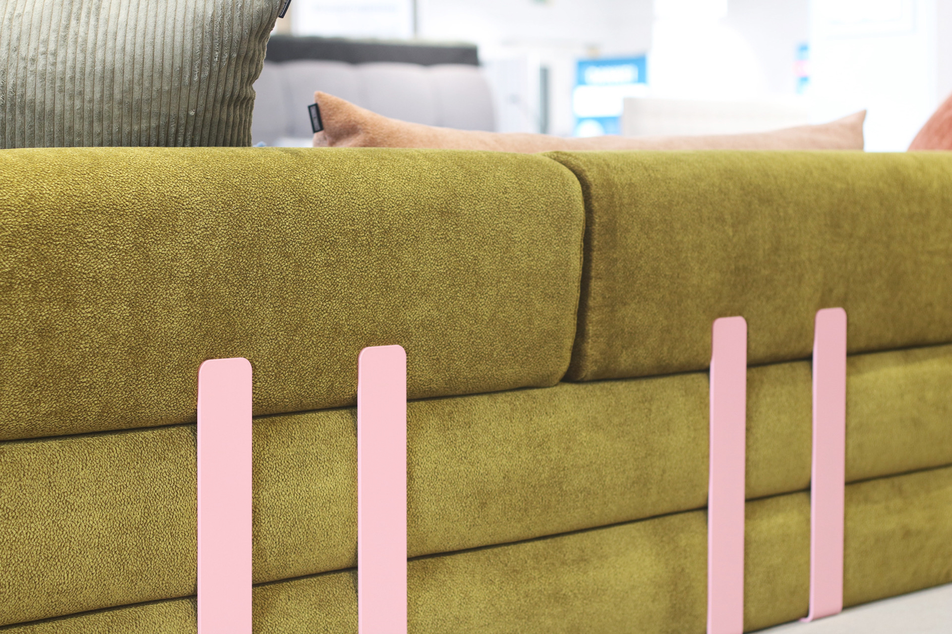 Nahaufnahme eines grün gepolsterten Sofas mit rosafarbenen vertikalen Akzenten an der Rückenlehne, das mit grünen und beigen Kissen ausgestattet ist. Die Szenerie erinnert an die Ausstellungsräume von Snooze Project vor dem verschwommenen Hintergrund eines modernen, hellen Raums.