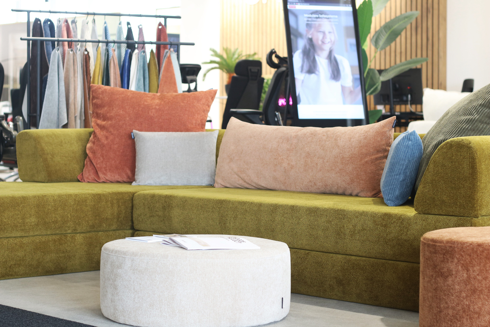 Ein modernes Wohnzimmer mit einem olivgrünen Sektionssofa, verschiedenen Kissen in Orange, Beige, Blau und Grau, einem runden beigen Ottomanen und einem Kleiderständer mit bunten Stoffen - die perfekte Inspiration für die Ausstellungsräume von Friends of Snooze Project.