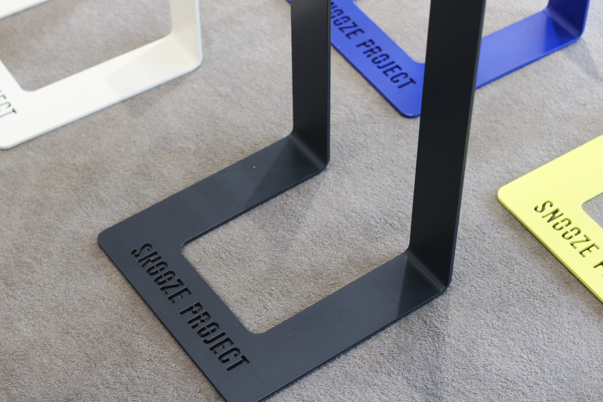 Die Metallständer in Schwarz, Weiß, Blau und Gelb sind auf einer grauen Oberfläche platziert. Auf jedem Ständer ist in der Nähe des Sockels der Schriftzug SNOOZE PROJECT ausgeschnitten, was sie zu perfekten Ausstellungsstücken für Snooze Project Showrooms macht.