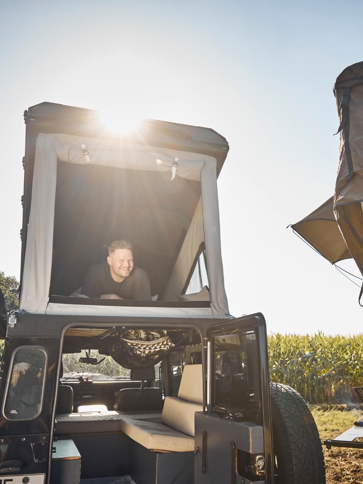 Eine Person lehnt sich lächelnd aus dem aufklappbaren Dachzelt eines Wohnmobils, während hinter ihr das Sonnenlicht scheint und im Hintergrund ein Feld mit hohen Pflanzen zu sehen ist.