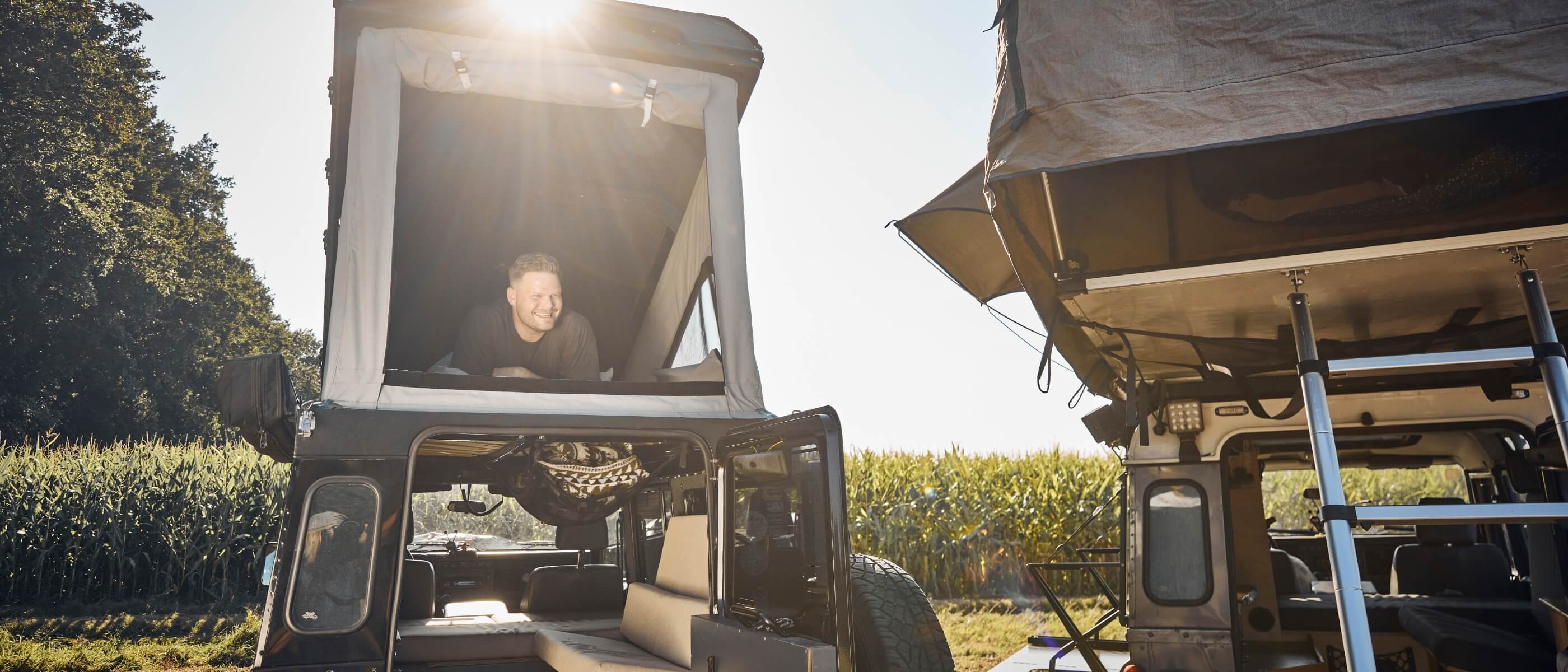 Eine Person lächelt aus einem Zelt auf dem Dach eines Wohnmobils, in dessen Nähe sich ein weiteres ähnliches Zelt und ein offenes Fahrzeug befinden. Die Sonne scheint hell über ein Feld mit hohen grünen Pflanzen im Hintergrund.