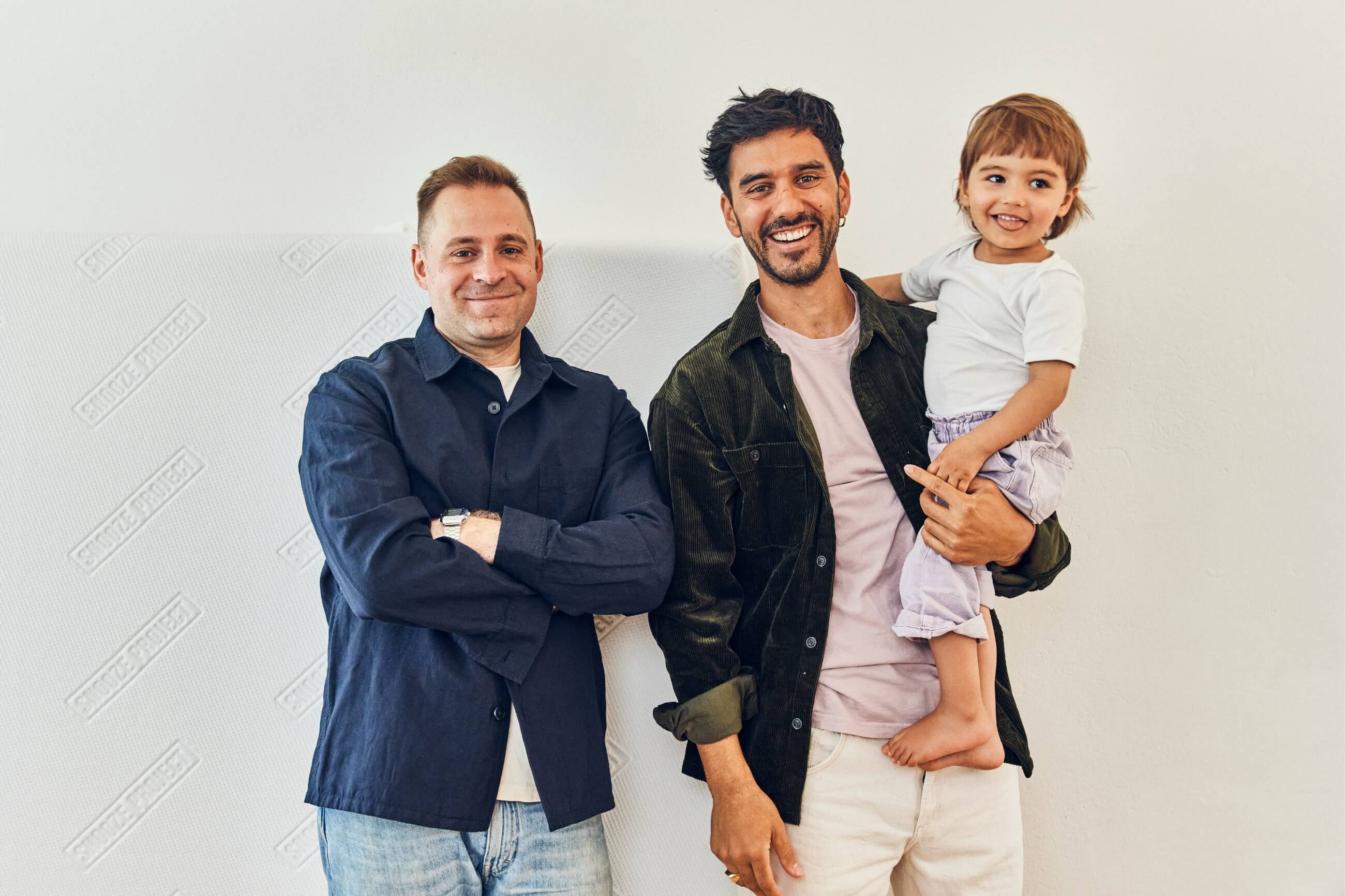 Zwei lächelnde Männer stehen Seite an Seite vor einer weißen Wand. Der eine hat die Arme verschränkt, der andere hält ein kleines Kind, das ebenfalls lächelt. Sie wirken alle glücklich und entspannt.