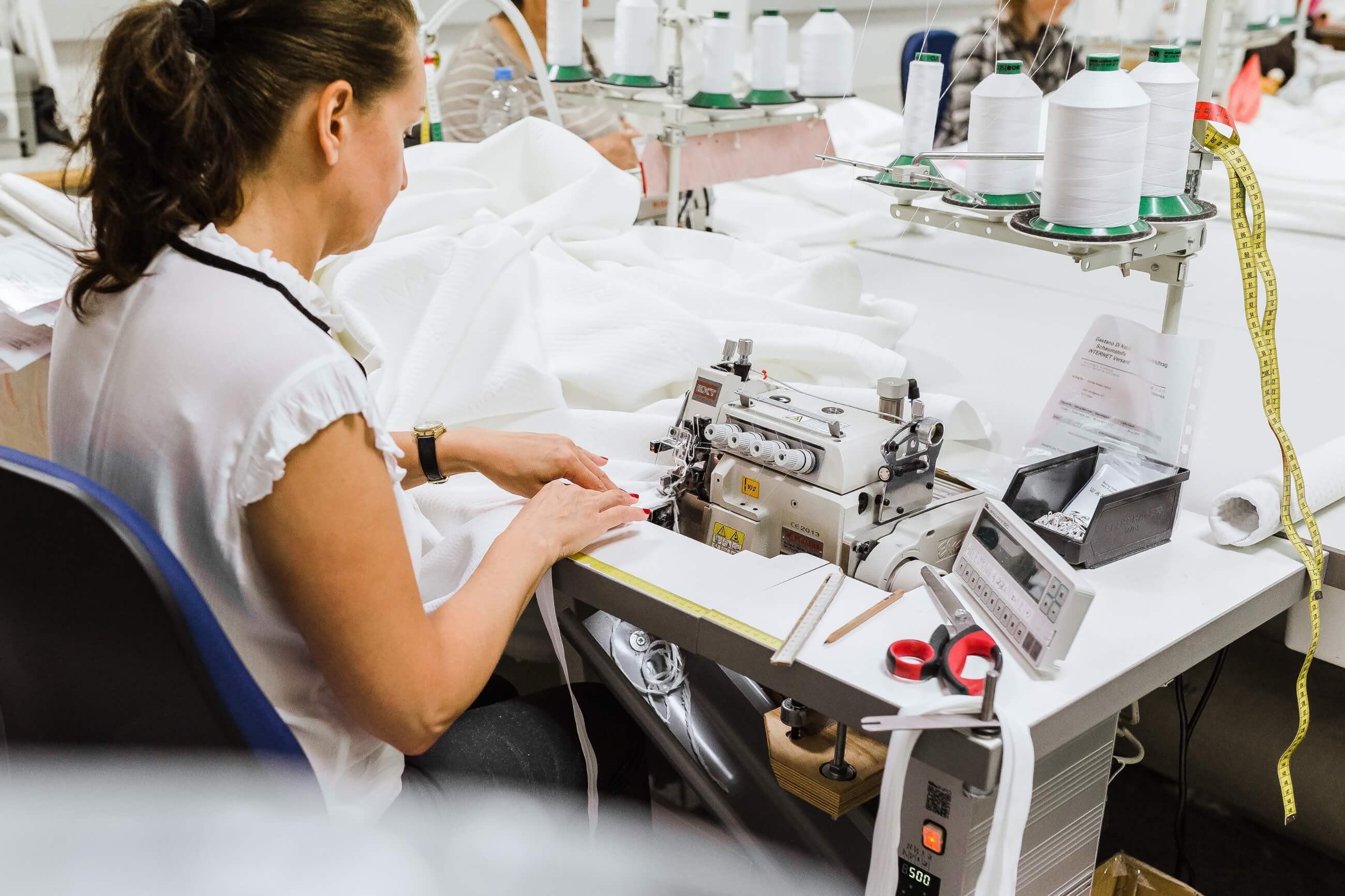 Eine Frau bedient eine Nähmaschine an einem Arbeitstisch, der mit weißem Stoff, Garnspulen, Scheren und anderen Nähwerkzeugen in einer Textilwerkstatt bedeckt ist.