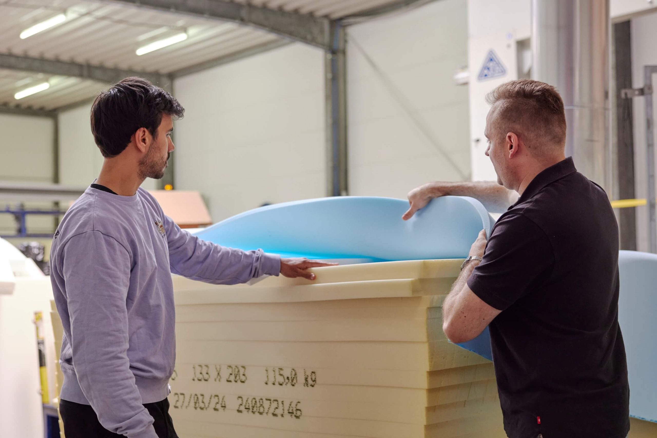 Zwei Männer inspizieren Schaumstoffplatten in einer industriellen Umgebung. Der eine hebt eine blaue Schaumstoffschicht auf einen Stapel gelben Schaumstoffs, während der andere beobachtet. Beide stehen in einer gut beleuchteten Werkstatt mit freiliegenden Balken und Geräten.