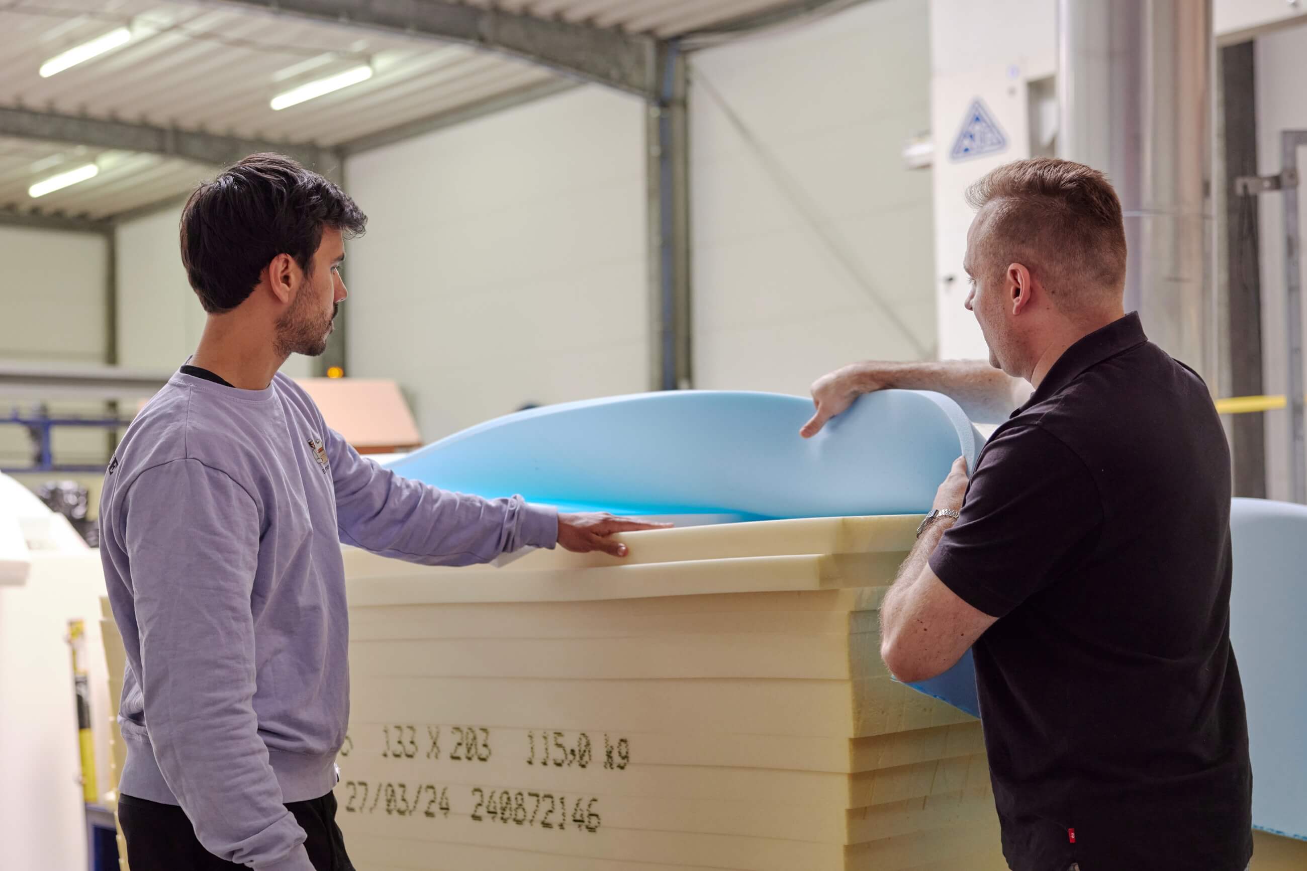 Zwei Männer inspizieren Schaumstoffplatten in einer industriellen Umgebung. Der eine hebt eine blaue Schaumstoffschicht auf einen Stapel gelben Schaumstoffs, während der andere beobachtet. Beide stehen in einer gut beleuchteten Werkstatt mit freiliegenden Balken und Geräten.