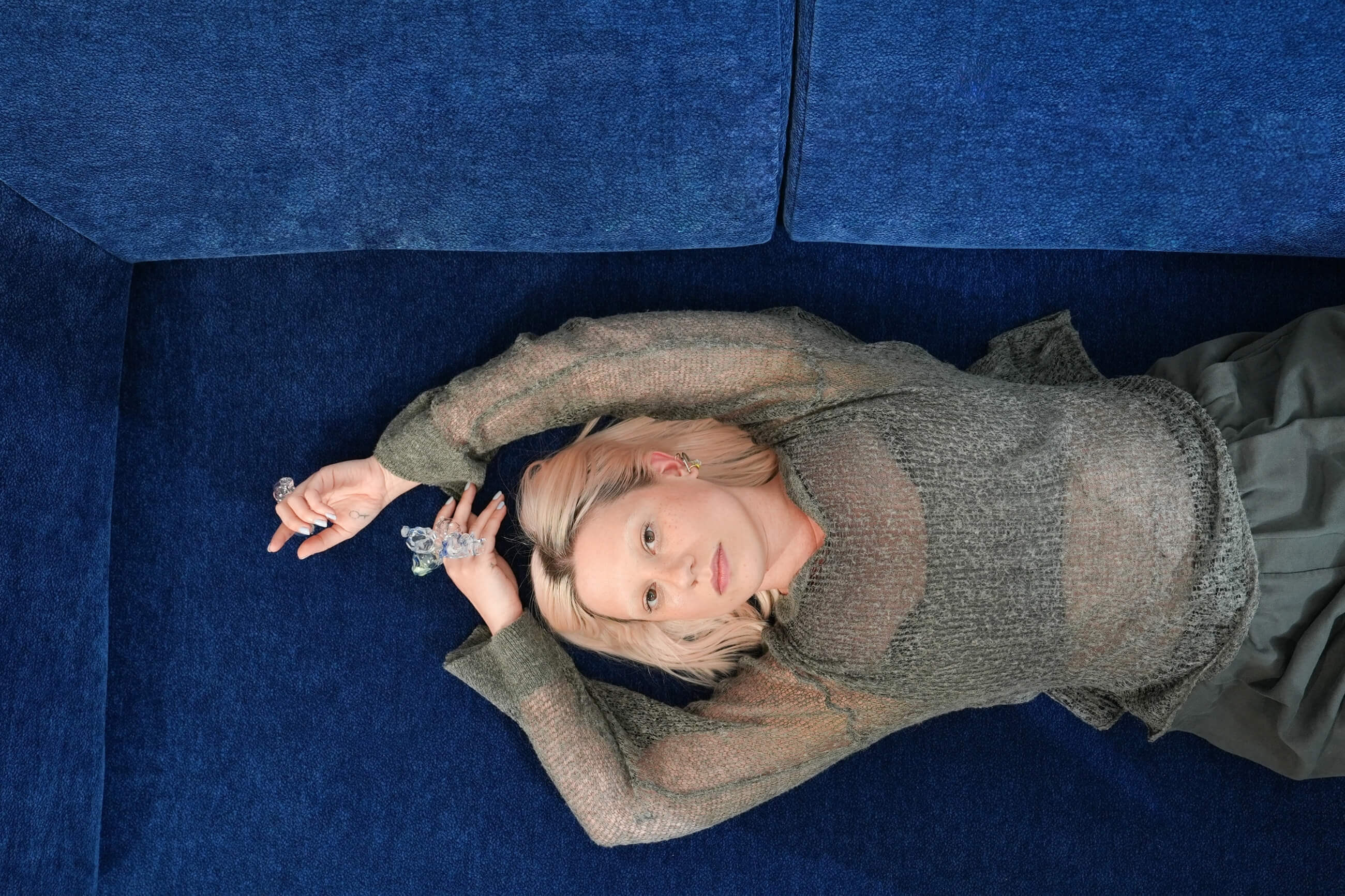 Eine Person mit hellem Haar liegt auf einem blauen Sofa und trägt ein durchsichtiges graues Oberteil und einen dunklen Rock. Ein Arm ist über ihren Kopf gestreckt, der andere ist angewinkelt und hält kleine dekorative Gegenstände in der Hand.