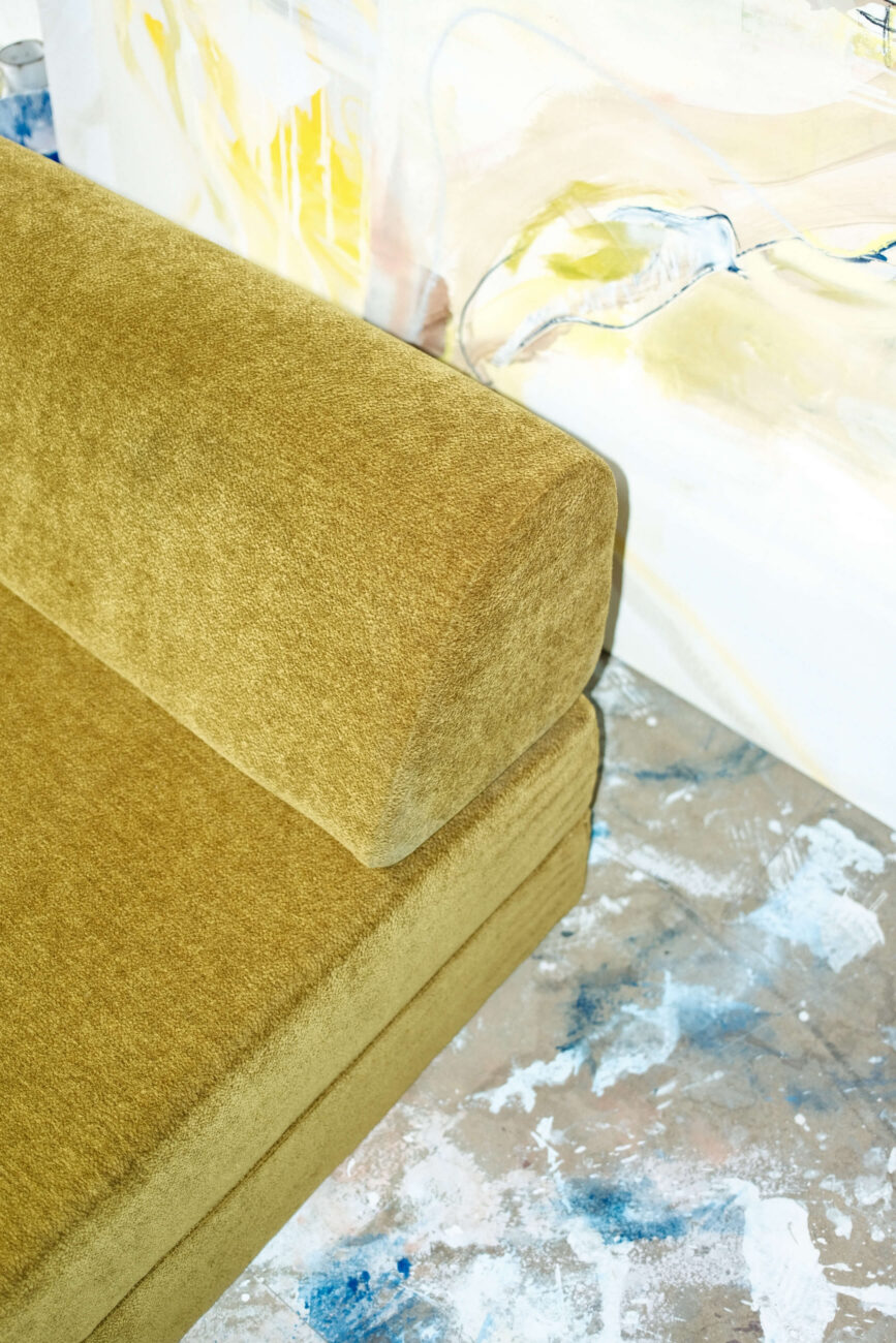 Nahaufnahme eines senfgelben Sofas mit rechteckiger Armlehne, das neben einer Wand mit einem abstrakten Gemälde und einem strukturierten Boden mit blauen und weißen Farbspritzern steht.
