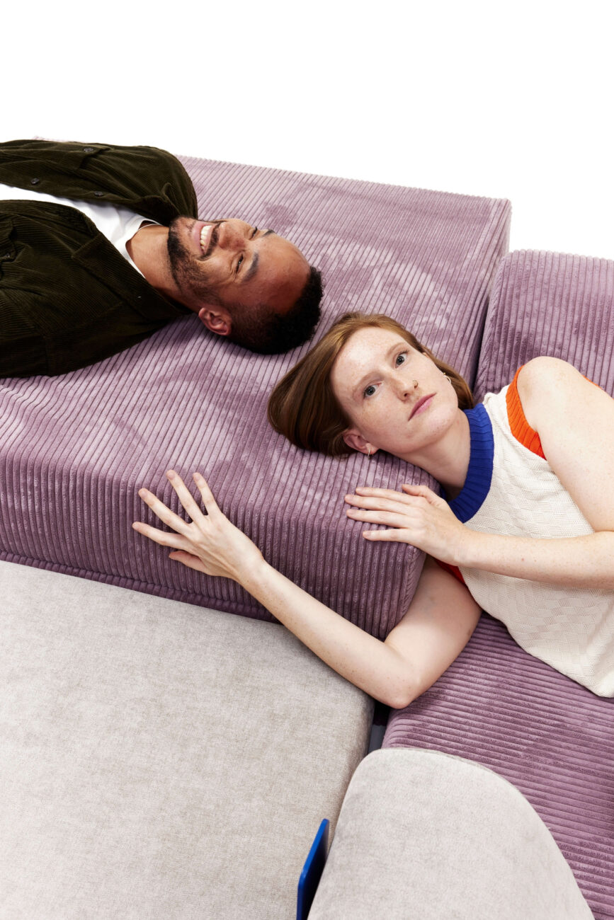 Ein Mann und eine Frau liegen nebeneinander auf pastellfarbenen Sofakissen mit Cordstruktur, wobei die Frau ihren Kopf und ihren Arm auf ein lavendelfarbenes Kissen vor einem weißen Hintergrund stützt.