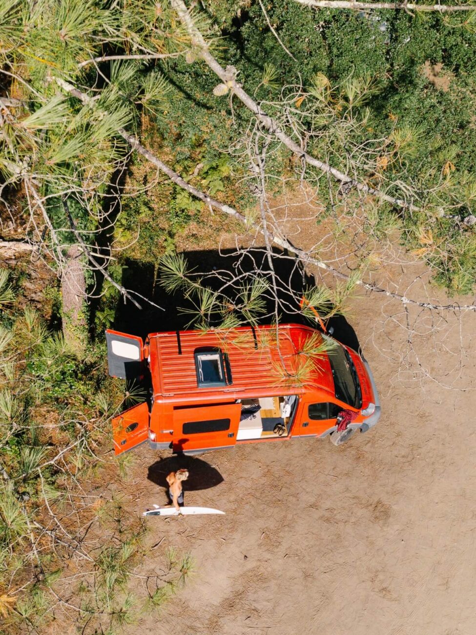 Overhead-Drohnenaufnahme eines orangefarbenen Wohnmobils mit offenen Türen. Eine Person steht mit einem Surfbrett neben dem Van, umgeben von Bäumen und trockenem Gestrüpp.