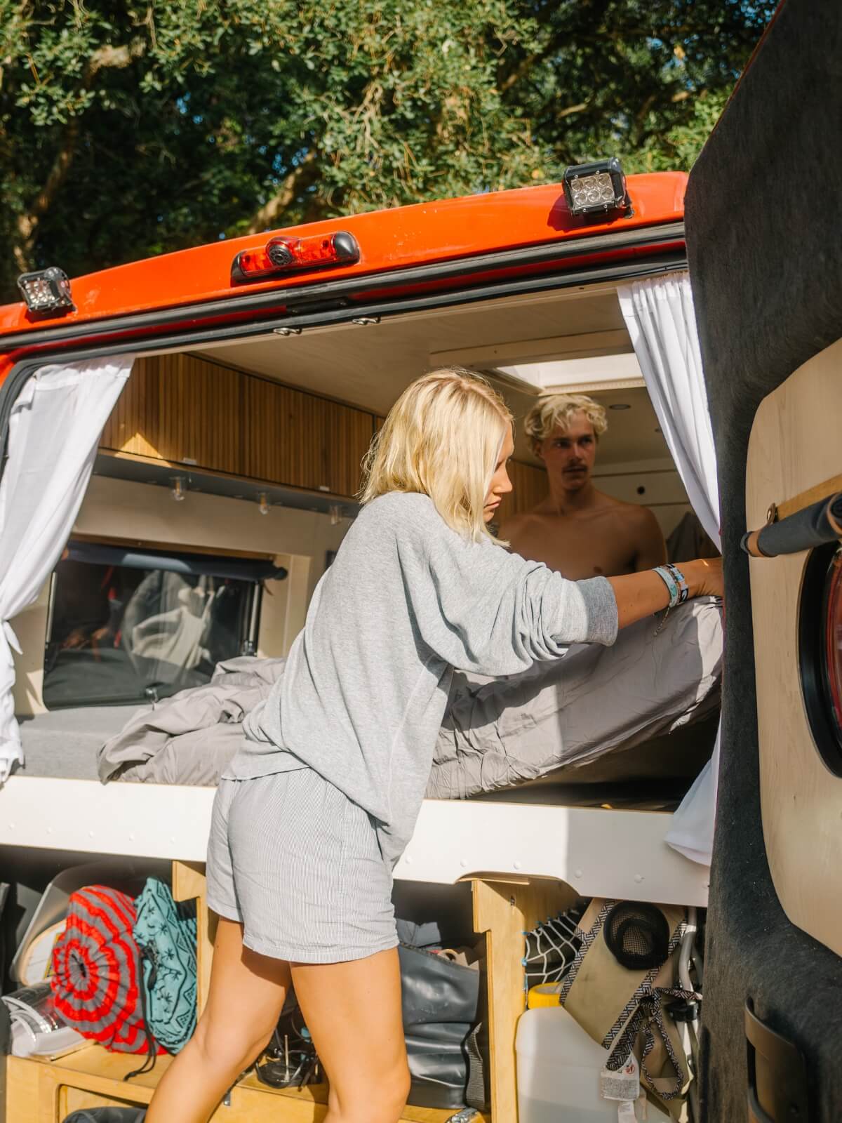 Eine Frau mit blondem Haar macht ein Bett in einem Wohnmobil, während ein Mann ohne Hemd mit blondem Haar von hinten zuschaut.