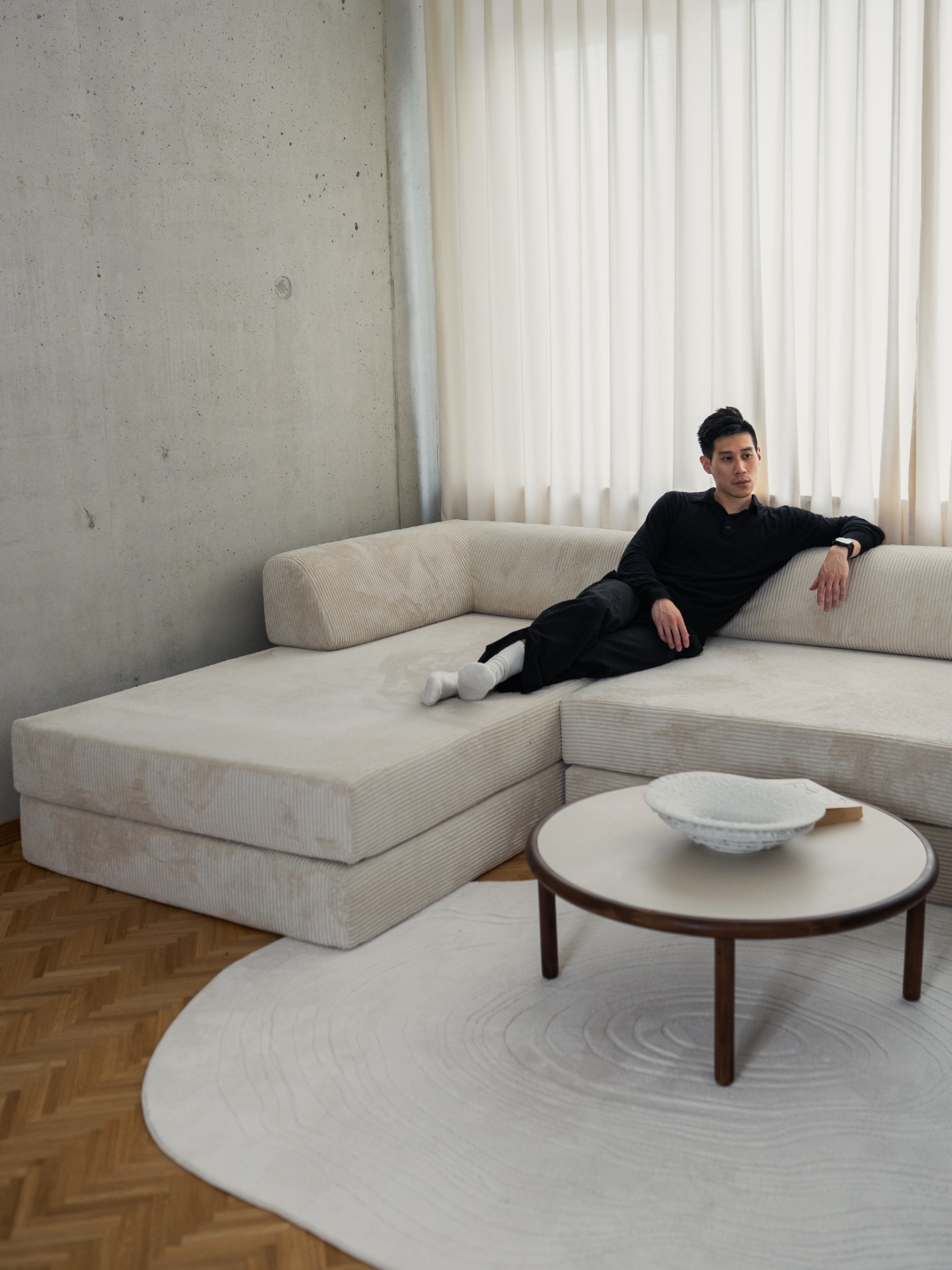 Ein Mann in dunkler Kleidung und weißen Socken macht es sich auf einem großen, cremefarbenen Ecksofa in einem modernen, minimalistischen Wohnzimmer gemütlich, das in neutralen Farbtönen gehalten ist und mit transparenten Vorhängen sowie einem runden Couchtisch auf einem runden Teppich ausgestattet ist.