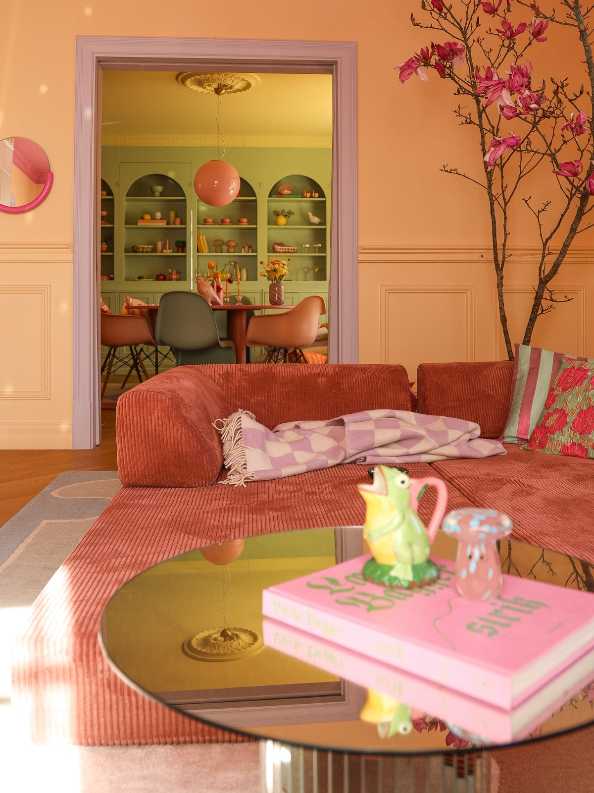 Ein gemütliches Wohnzimmer mit einem rosa Sofa, einer karierten Decke und einem Couchtisch, auf dem rosa Bücher und eine Tasse in Froschform stehen. Im Hintergrund ist durch eine Türöffnung ein pastellgrüner Essbereich mit Regalen und einem runden Tisch zu sehen.