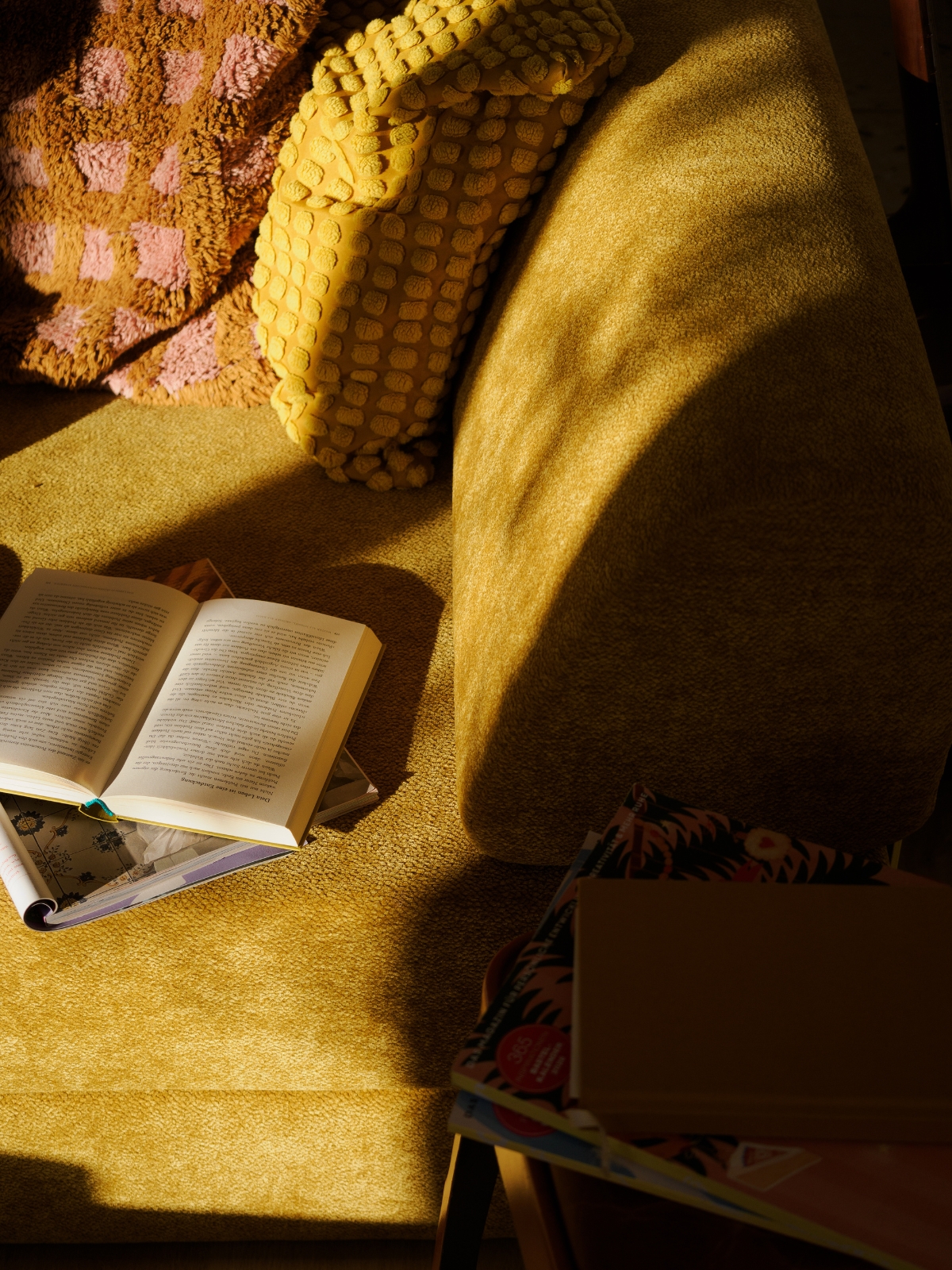 Ein sonnengelb leuchtendes Sofa mit einem gelben Kissen mit Struktur und einer rosa Decke. Auf der Sitzfläche liegt ein aufgeschlagenes Buch, daneben auf einer Holzfläche ein geschlossenes Notizbuch und eine bunte Mappe.