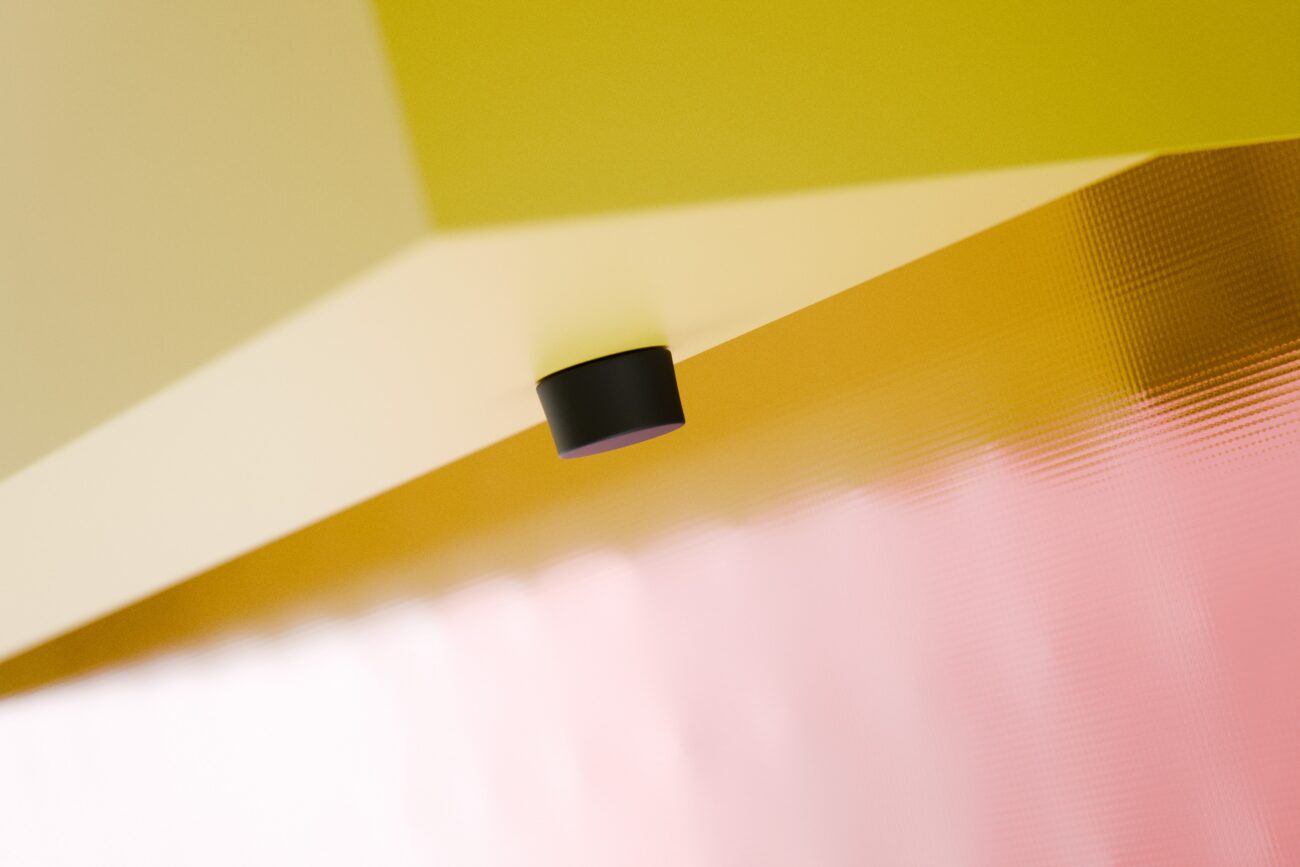 An der Unterseite einer geometrischen gelben Fläche, über einer strukturierten, durchscheinenden rosa Platte, ist ein kleiner schwarzer Zylinder befestigt. Die Komposition ist minimalistisch und abstrakt.