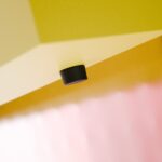 An der Unterseite einer geometrischen gelben Fläche, über einer strukturierten, durchscheinenden rosa Platte, ist ein kleiner schwarzer Zylinder befestigt. Die Komposition ist minimalistisch und abstrakt.