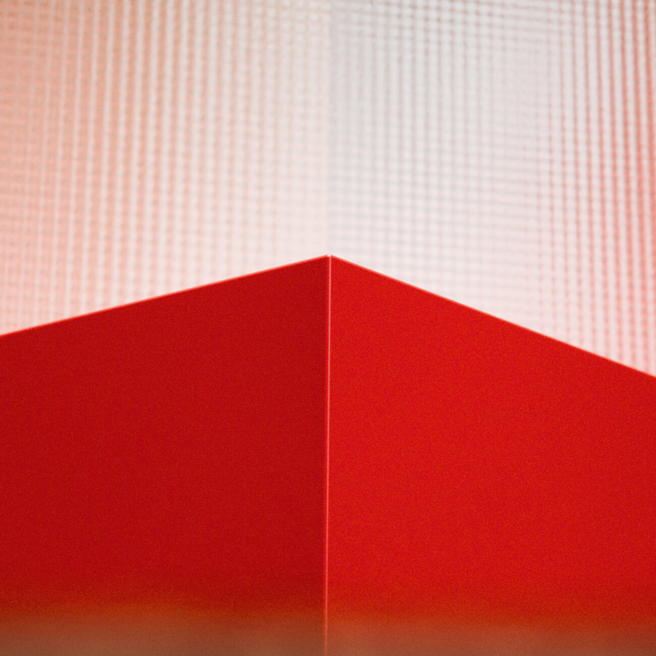 LUCA001-mood_img003 Eine rote geometrische Form mit einer scharfen Ecke ist nach oben gerichtet und hebt sich von einem strukturierten, hellrosa Hintergrund ab. Das Bild zeichnet sich durch kräftige Linien und eine abstrakte, minimalistische Komposition aus.