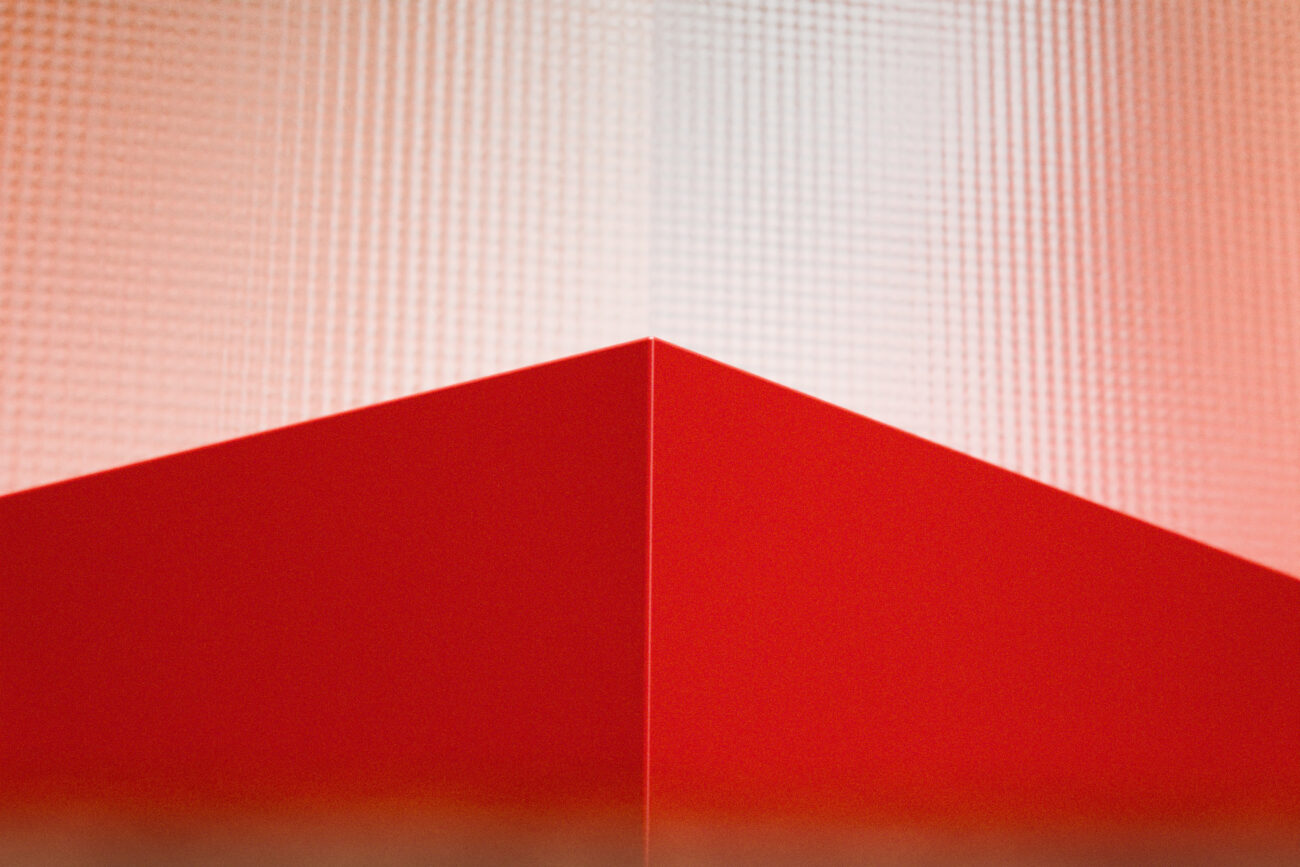 Eine rote geometrische Form mit einer scharfen Ecke ist nach oben gerichtet und hebt sich von einem strukturierten, hellrosa Hintergrund ab. Das Bild zeichnet sich durch kräftige Linien und eine abstrakte, minimalistische Komposition aus.
