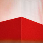 Eine rote geometrische Form mit einer scharfen Ecke ist nach oben gerichtet und hebt sich von einem strukturierten, hellrosa Hintergrund ab. Das Bild zeichnet sich durch kräftige Linien und eine abstrakte, minimalistische Komposition aus.