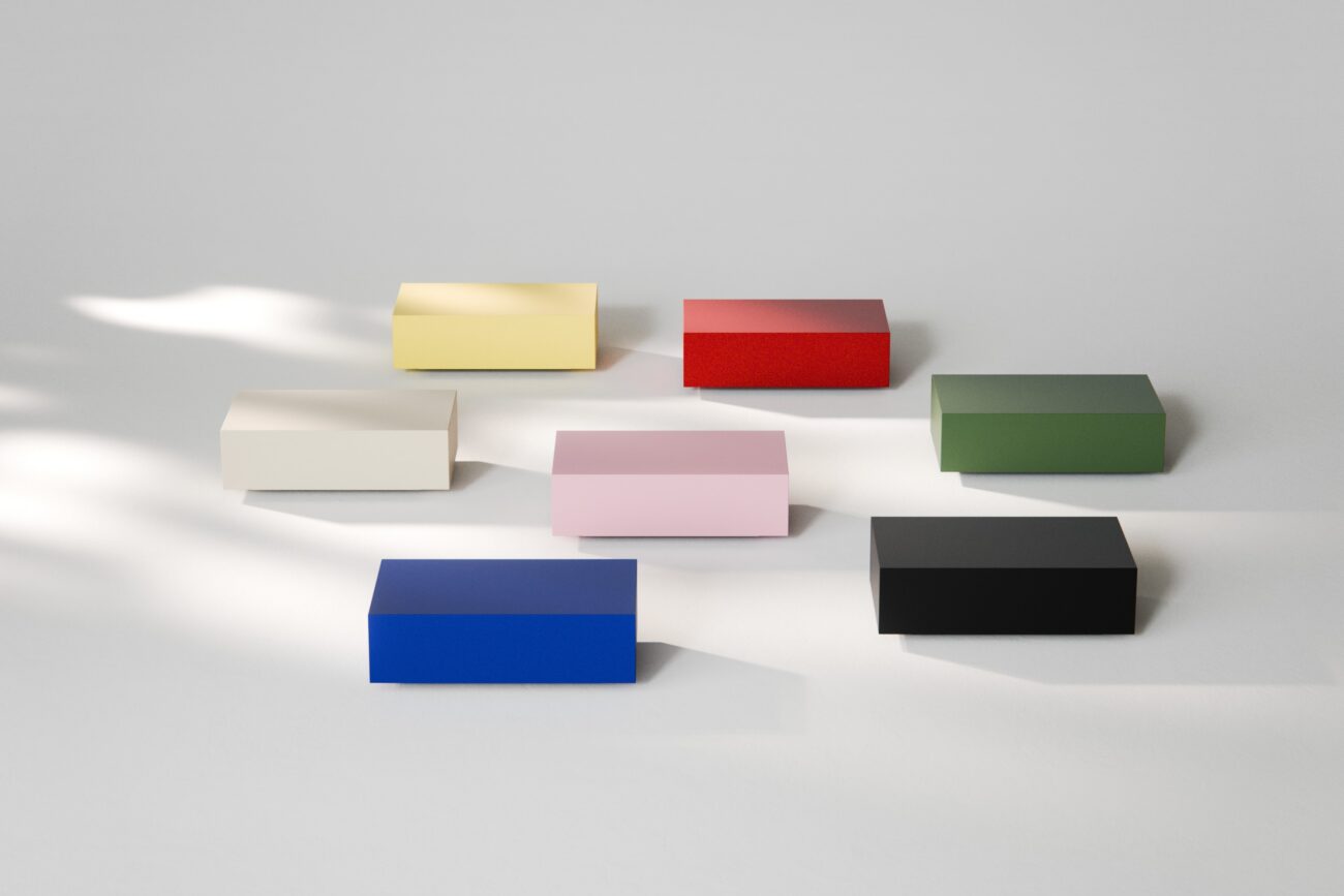 Sieben kleine, rechteckige Schachteln in verschiedenen Farben – gelb, rot, grün, schwarz, blau, rosa und cremefarben – sind auf einer glatten, hellgrauen Fläche mit sanften Schatten und hellen Flecken angeordnet.