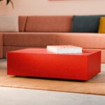 Ein modernes Wohnzimmer mit einem roten, rechteckigen Couchtisch, auf dem ein geschlossenes Buch liegt, darunter ein beiger Teppich und im Hintergrund ein hellbraunes Ecksofa mit bunten Kissen.