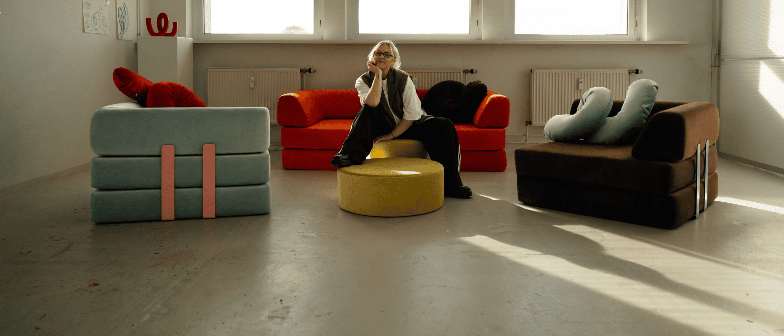 Eine Person mit hellem Haar und Brille sitzt auf einem gelben Hocker in einem modernen Raum, umgeben von bunten, ungewöhnlich geformten Sofas und großen Fenstern, durch die Sonnenlicht hereinströmt.