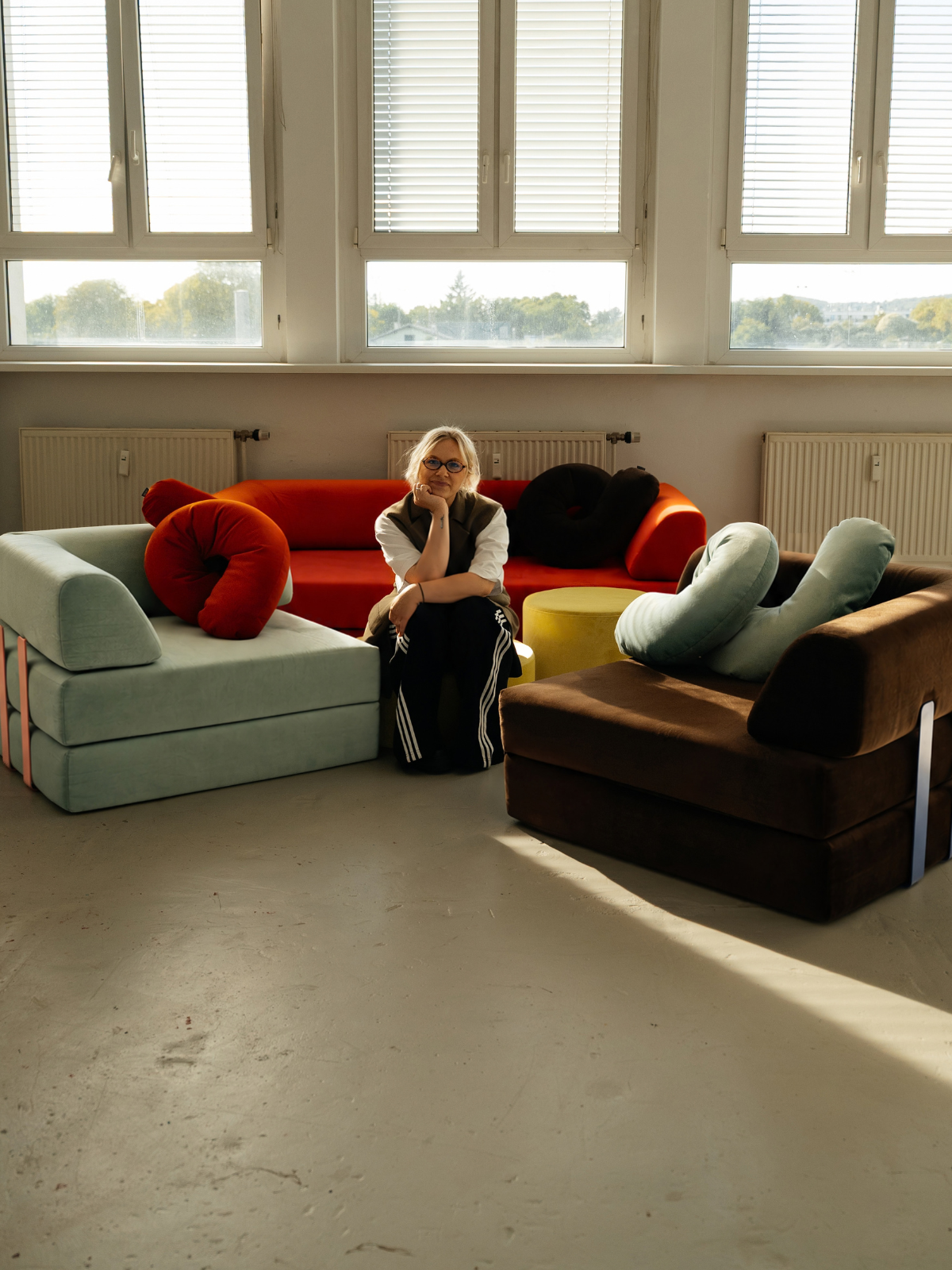 Eine Person sitzt auf dem Boden, umgeben von bunten, einzigartig geformten Sofas unter großen Fenstern, durch die Tageslicht hereinströmt.