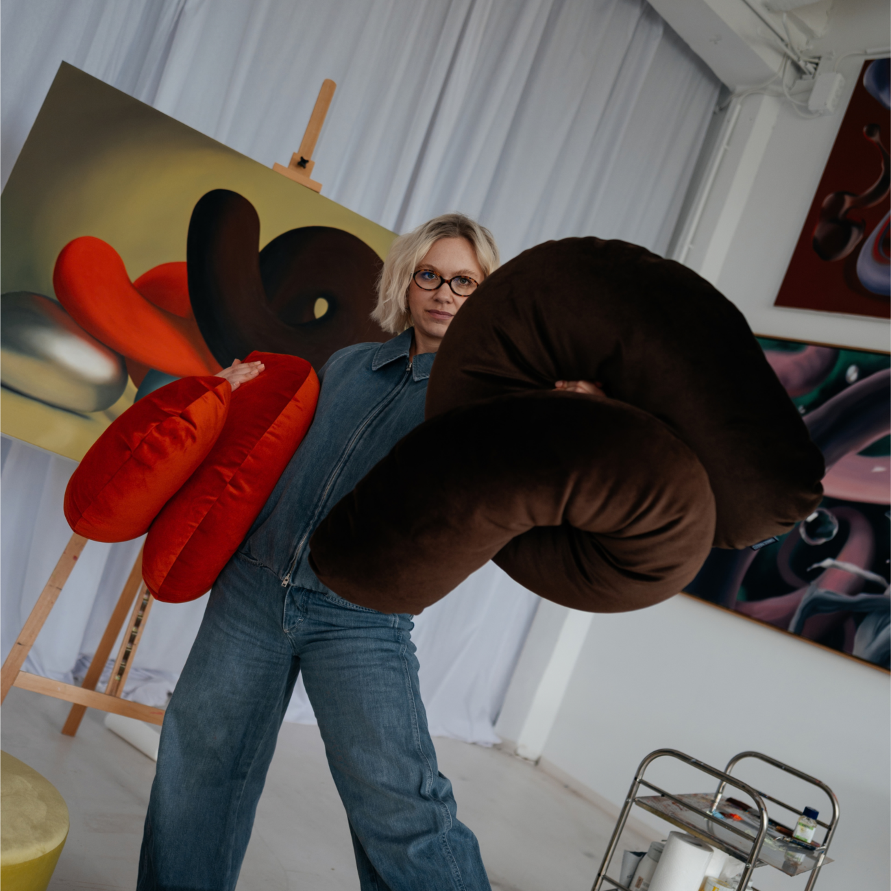 SPKJU20x260-SS-Mood_imgG-1 Eine Person in einem Jeans-Overall hält große, flauschige SP x Ju Schnee Deko-Kissen in einem hellen Atelier, im Hintergrund sind abstrakte Gemälde von Ju Schnee zu sehen.