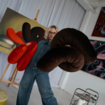 Eine Person in einem Jeans-Overall hält große, flauschige SP x Ju Schnee Deko-Kissen in einem hellen Atelier, im Hintergrund sind abstrakte Gemälde von Ju Schnee zu sehen.