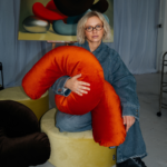 Eine Frau mit blonden Haaren und Brille sitzt auf einem gelben Ottoman und hält ein geschwungenes SP x Ju Schnee-Dekokissen in der Hand. Sie trägt blaue Jeans, im Hintergrund sind abstrakte Kunstwerke von Ju Schnee zu sehen.