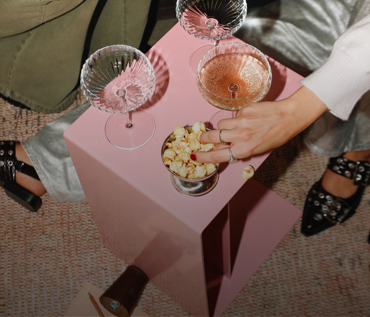 Eine Hand greift in eine kleine Schüssel mit Popcorn, die auf einem rosa Beistelltisch steht, auf dem drei Cocktailgläser stehen.