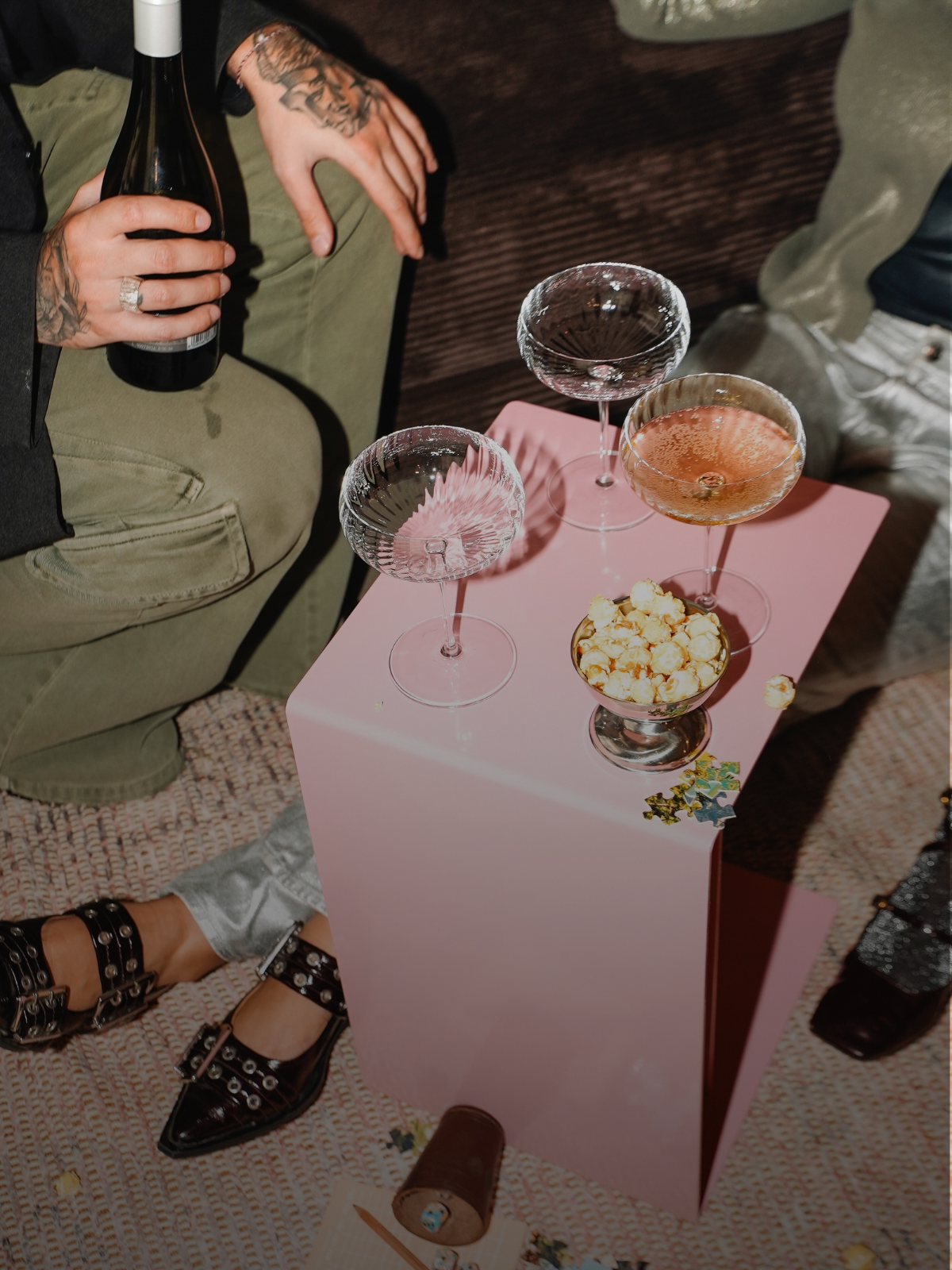 Zwei Personen sitzen auf einem Sofa, auf einem rosa Beistelltisch stehen eine Flasche Wein, zwei Coupé-Gläser und eine Schüssel Popcorn.