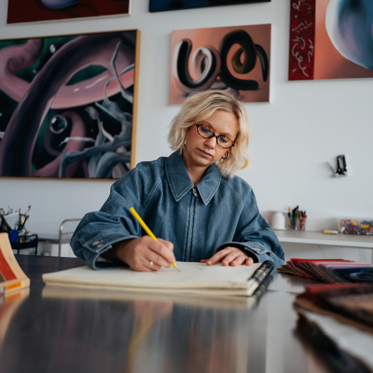 Eine Person mit blonden Haaren und Brille zeichnet in einem großen Notizbuch an einem Tisch in Jus Kunstatelier, umgeben von abstrakten Gemälden an der Wand.