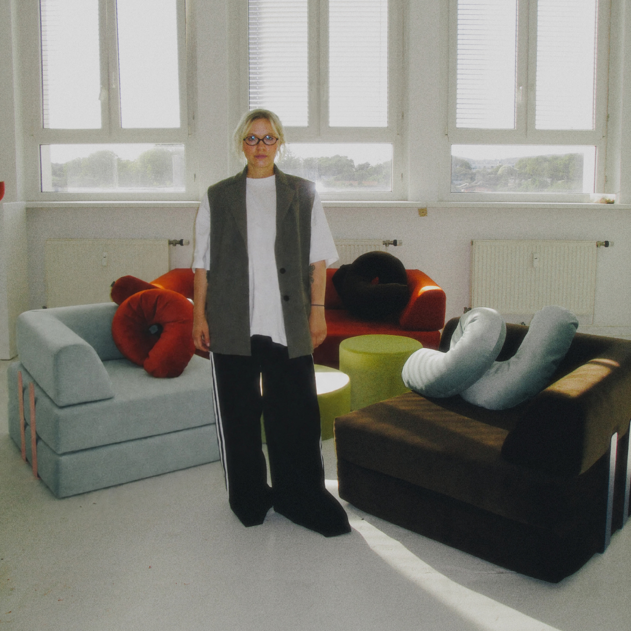 Eine Person mit Brille steht in einem hellen Raum mit großen Fenstern, umgeben von farbenfrohen, modernen Sofas und Ju-Schnee-Kissen, und schafft so eine lebendige Erlebniswelt aus einzigartigen, kissenartigen Dekorationselementen.