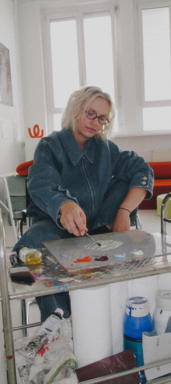 Eine Person mit Brille und blonden Haaren, gekleidet in ein blaues Jeansoutfit, sitzt in einer hell beleuchteten Erlebniswelt und mischt Farbe auf einer Palette, umgeben von Malutensilien und einem Ju Schnee-Kissen.
