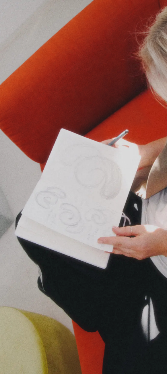 Eine Person sitzt auf einem roten Stuhl und skizziert mit Bleistift spiralförmige Muster in ein weißes Notizbuch – ganz im Stil von Ju Schnee. Zu sehen sind nur ihre Hände, ein Teil ihres Oberkörpers und das Skizzenbuch.